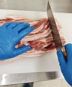 厨房の白いまな板の上で、青いニトリル手袋を着用し、大きな中華包丁で生の豚バラ肉のスライスを重ねて切る調理の様子。ステンレス製の調理台。