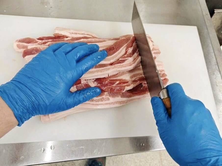 厨房の白いまな板の上で、青いニトリル手袋を着用し、大きな中華包丁で生の豚バラ肉のスライスを重ねて切る調理の様子。ステンレス製の調理台。